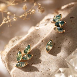 May include: Three gold-toned stud earrings featuring marquise-cut moss agate stones. One earring is a cluster of three stones, another is a cluster of two stones, and the third is a single marquise-cut stone, all displaying green inclusions.