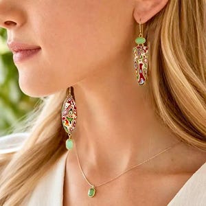 Pode incluir: Um par de brincos pendentes dourados e um colar a condizer. Os brincos apresentam um pendente triangular com um padrão de redemoinho multicolorido e uma pequena pedra verde. O colar tem um pendente oval com um padrão semelhante.