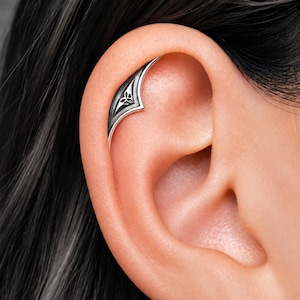 Può includere: Un ear cuff color argento dal design curvo e appuntito, con finitura ossidata e un piccolo simbolo triquetra inciso. Il gioiello è posizionato lungo la cartilagine superiore dell'orecchio, evidenziando una texture metallica e bordi netti.