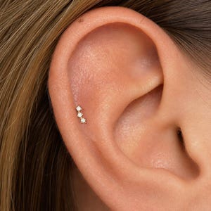 May include: A gold-toned ear cartilage piercing stud featuring three small, clear, round-cut stones arranged in a vertical, slightly curved line. The jewelry is positioned along the upper edge of the ear, secured with a thin metal post.