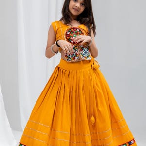 May include: A bright yellow two-piece outfit consisting of a sleeveless crop top and a long, pleated skirt. The top features a circular embroidered patch with mirrors and colourful threadwork. The skirt has horizontal gold trim and a patterned border.