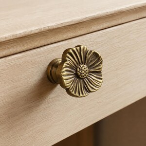 May include: A brass-toned metal drawer knob shaped like a five-petaled flower with a textured center. The knob features a metallic finish with darkened recessed areas between the petals and is attached to a light-colored wooden drawer front.
