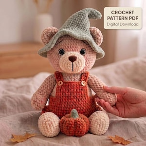 May include: A crocheted teddy bear wearing a sage green pointed hat and orange overalls with two small buttons. The bear has dark eyes and a brown nose. A small crocheted pumpkin sits between its feet. Text reads: CROCHET PATTERN PDF Digital Download.