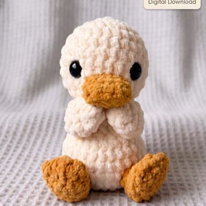 Puede incluir: Un patito de peluche tejido a ganchillo en color crema con un acabado de hilo texturizado, que presenta un pico y patas de color naranja brillante, y dos grandes ojos de seguridad negros. Una etiqueta muestra CROCHET PATTERN PDF Digital Download.