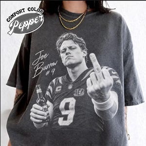 Peut inclure: Un t-shirt gris anthracite à manches courtes avec un graphique en noir et blanc de Joe Burrow portant un maillot numéro 9, tenant une bouteille et faisant un doigt d'honneur. Le texte indique Joe Burrow #9 et Comfort Color Pepper.