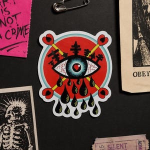 Puede incluir: Una pegatina que muestra un ojo azul central con una pupila en forma de corazón, sobre un fondo circular rojo. Dos flechas cruzadas con extremos en forma de corazón atraviesan el ojo, mientras caen lágrimas negras. El diseño incluye siluetas negras de árboles sobre el ojo.