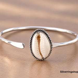 Peut inclure: Un bracelet manchette en argent avec un coquillage cauri ovale central serti dans une bordure en métal torsadé. Le bracelet présente un design ouvert avec des bandes en argent poli et lisse qui s'affinent aux extrémités. Le texte Silverringsshop26 est visible.