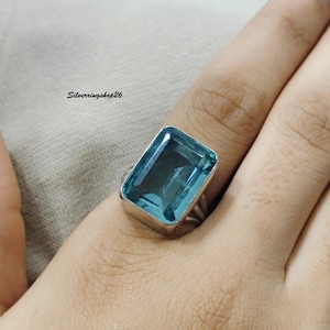 Puede incluir: Un anillo de plata con una gran piedra azul rectangular facetada engastada en un bisel de metal liso. La banda tiene un diseño dividido y el texto Silverringshop26 es visible en la esquina superior izquierda de la imagen.