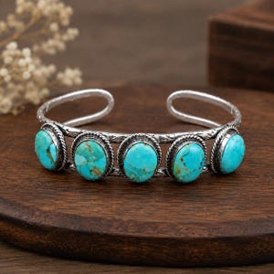 May include: A silver cuff bracelet featuring five oval-shaped turquoise stones with brown matrix patterns. The metal band has a textured, hammered finish and a twisted rope border surrounding each stone, creating a detailed, open-ended design.