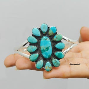 May include: A silver cuff bracelet featuring a large central oval turquoise stone surrounded by twelve smaller teardrop-shaped turquoise stones. The metal setting has a dark oxidized finish between the stones. The text Silverringshop26 is visible.