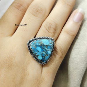 Peut inclure: Une bague triangulaire ornée d'une pierre bleue vibrante avec des motifs de matrice bruns complexes. La pierre est sertie dans une monture en argent poli avec un détail de bordure en corde torsadée, créant un aspect texturé sur l'anneau.