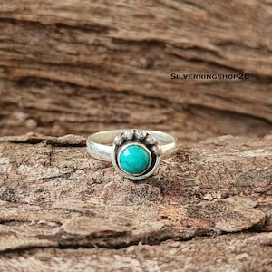 Peut inclure: Une bague en argent avec une pierre turquoise ronde et vive sertie dans une monture. Le sertissage est orné d'une série de petites sphères en argent en relief le long du bord supérieur. Le texte SILVERRINGSHOP26 est visible en haut à droite.
