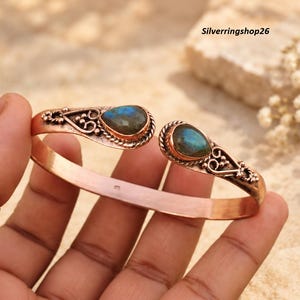 May include: A copper cuff bracelet featuring two teardrop-shaped labradorite gemstones at the open ends. The metal band displays intricate scrollwork and beaded detailing surrounding each stone. The text Silverringshop26 is visible.