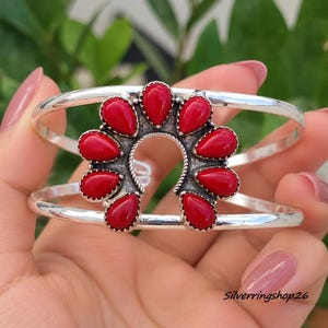 May include: A silver cuff bracelet featuring a horseshoe-shaped centrepiece adorned with nine teardrop-shaped red stones. The metal has a polished finish with oxidised detailing around the stone settings. The text Silverringshop26 is visible.