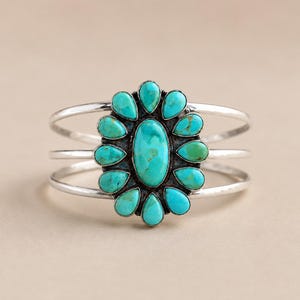 May include: A silver cuff bracelet featuring a large, central oval turquoise stone surrounded by twelve smaller, teardrop-shaped turquoise stones arranged in a floral pattern. The bracelet band consists of three thin, polished silver wires.