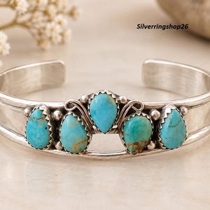 May include: A silver cuff bracelet featuring five teardrop-shaped turquoise stones set in serrated bezels. The centre stone is slightly elevated, flanked by two pairs of smaller stones. The metal band has a smooth, polished finish. Text reads Silverringshop26.