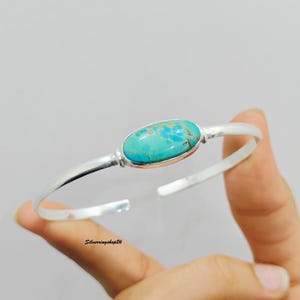 May include: A silver cuff bracelet featuring a central oval-shaped turquoise stone with natural brown matrix veining. The polished metal band has an open design, and the text "Silverringshop26" is printed in small black letters near the bottom edge.