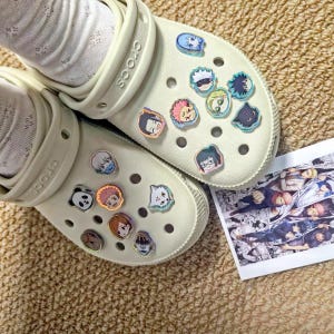 May include: A pair of cream-coloured clogs featuring multiple small, colourful anime-style character charms inserted into the ventilation holes. The clogs have a textured heel strap and the brand name Crocs embossed on the side, accompanied by a small rectangular print showing a black and white manga panel.