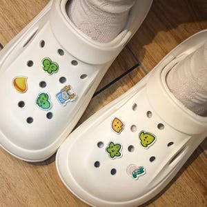 Puede incluir: Un par de zuecos de espuma color blanco roto con múltiples orificios de ventilación. Los zapatos están decorados con varios dijes pequeños y coloridos, incluyendo hojas verdes, una gota amarilla, una criatura azul con astas y otras formas.
