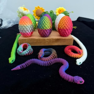 Puede incluir: Una colección de serpientes articuladas y huevos de dragón impresos en 3D con patrones de escamas texturizados. Los huevos presentan colores degradados que incluyen rosa, naranja, verde, azul y morado. Las serpientes son multicolores.