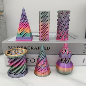 May include: A collection of six 3D printed objects featuring a gradient of purple, pink, orange, and green colours. The shapes include a pyramid, a rectangular prism, a cylindrical lattice, a spool, a cone, and a rounded bulbous form with cutouts.