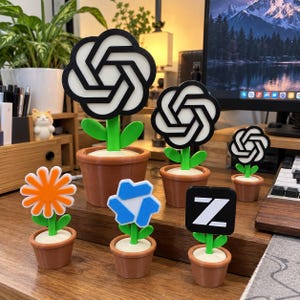 Könnte beinhalten: Eine Sammlung von sechs kleinen künstlichen Topfblumen mit verschiedenen Logos und Formen. Drei Blumen zeigen das schwarz-weiße OpenAI-Logo, eine ist eine leuchtend orangefarbene Margerite, eine hat eine blaue geometrische Form und eine zeigt ein weißes Z auf einem schwarzen Quadrat.