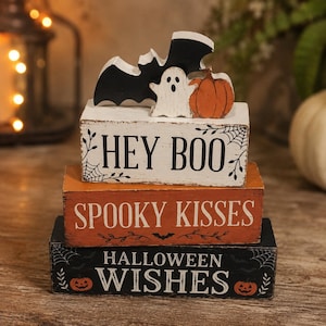 Può includere: Una pila di tre blocchi rettangolari in legno con bordi usurati. Il blocco bianco superiore presenta un pipistrello nero, un fantasma bianco, una zucca arancione e il testo HEY BOO. Il blocco arancione centrale riporta SPOOKY KISSES. Il blocco nero inferiore riporta HALLOWEEN WISHES.