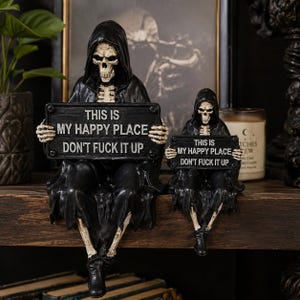 May include: Two black-robed skeleton figurines sitting side-by-side, each holding a rectangular black sign with white text that reads: THIS IS MY HAPPY PLACE DON'T FUCK IT UP. The skeletons have visible rib cages, skulls, and wear black hooded cloaks.