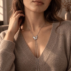 Op de afbeelding: Een zilverkleurige lariat-ketting met een vlinderhanger met iriserende witte inleg. De fijne ketting loopt door de vlinder, waardoor twee lange, slanke strengen ontstaan die verticaal vanaf het midden van de borst naar beneden hangen.