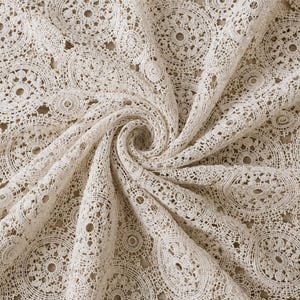 Pode incluir: Um tecido de renda em tom off-white com um padrão repetitivo de motivos circulares complexos. O têxtil está reunido numa espiral central, exibindo o design delicado e aberto, bem como a costura detalhada dos elementos redondos interligados.