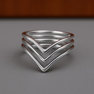 May include: A silver-coloured ring featuring a multi-layered chevron design. The piece consists of four distinct, smooth, polished bands that converge into a single V-shaped point at the centre, creating a stacked appearance with a metallic finish.