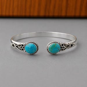 May include: A silver cuff bracelet featuring two oval-shaped turquoise stones at the open ends. The metal band displays a twisted rope texture and intricate scrollwork detailing surrounding each stone, creating a balanced and symmetrical design.
