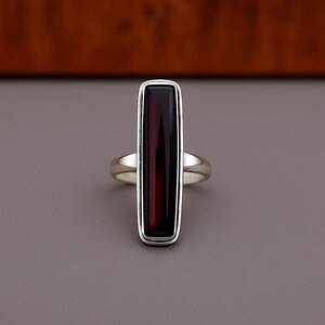 May include: A silver ring featuring a long, vertical, rectangular dark red gemstone set in a smooth metal frame. The stone has a deep, translucent appearance with a polished finish, and the band is a simple, polished silver metal loop.