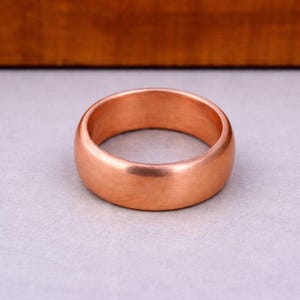 May include: A solid copper ring with a smooth, polished finish and a rounded band profile. The metal has a warm, reddish-brown metallic luster, showcasing a simple, continuous circular design without any engravings, stones, or additional decorative elements.
