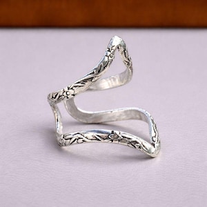 May include: A silver-toned metal ring featuring an open, wavy, and angular design. The surface of the band is decorated with a repeating embossed floral pattern consisting of small flowers and leaves along the entire length of the metal structure.