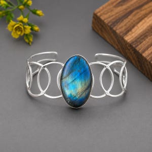 May include: A silver cuff bracelet featuring an oval-shaped labradorite gemstone with blue and gold iridescence. The band is constructed from a series of interconnected silver circles, creating an openwork design that wraps around the wrist.