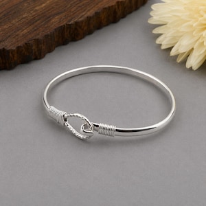 May include: A polished silver bangle bracelet featuring a hook-and-eye style clasp mechanism. The closure consists of a textured, hammered metal loop secured by two sections of fine wire wrapping on either side of the circular band.