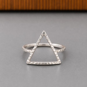 May include: A silver ring featuring a prominent open triangle shape as the centrepiece. The band and the triangular frame both exhibit a textured, hammered finish that creates a series of small, reflective indentations across the metal surface.
