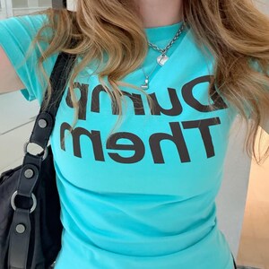 Puede incluir: Una camiseta de manga corta de color turquesa brillante con el texto "Dump Them" impreso en letras grandes, negritas y de color marrón oscuro en el pecho. La camiseta tiene cuello redondo y se combina con un bolso de hombro negro con herrajes plateados.