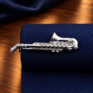 Op de afbeelding: Een zilverkleurige metalen dasclip in de vorm van een saxofoon. Het accessoire heeft een gepolijste, reflecterende afwerking met gedetailleerde gravures die de kleppen en de beker van het instrument weergeven, bevestigd op een donkerblauwe textuurstof.