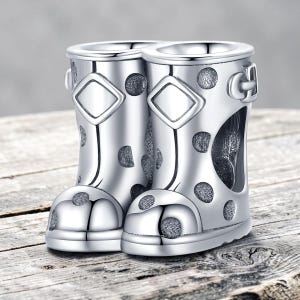 May include: A silver-toned charm shaped like a pair of rain boots featuring a polka dot pattern, a diamond-shaped detail on the front of each shaft, and a small buckle accent on the side of the right boot, all rendered in a polished metallic finish.