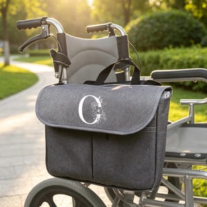 以下が含まれることがあります： チャコールグレーの車椅子用バッグ。フラップ部分には、花柄のラインアートで装飾された白い文字「O」がプリントされています。バッグには2つの黒いハンドルがあり、黒いストラップで金属製の車椅子のフレームに取り付けられています。