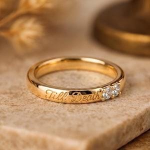 Könnte beinhalten: Ein goldener Ring mit der eingravierten Schreibschrift Till Death auf der Außenseite. Der Ring ist mit zwei kleinen, klaren, rund geschliffenen Steinen besetzt, die nebeneinander neben der Gravur liegen und eine polierte Metalloberfläche aufweisen.