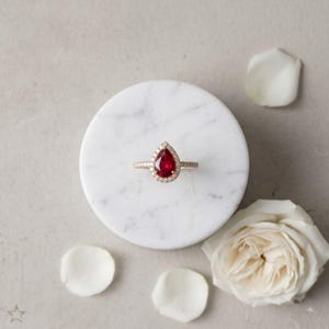 Pode incluir: Um anel com uma pedra preciosa vermelha em forma de pera, rodeada por um halo de pequenas pedras brancas. A banda é fina e metálica, centrada numa superfície circular de mármore branco, destacando o contraste da pedra vermelha intensa.