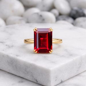 Könnte beinhalten: Ein Goldring mit einem großen, rechteckigen, im Smaragdschliff geschliffenen tiefroten Edelstein, der von vier Krappen gehalten wird. Die Ringschiene ist glatt und poliert und unterstreicht mit ihrem schlichten Design die leuchtende Farbe des Steins.