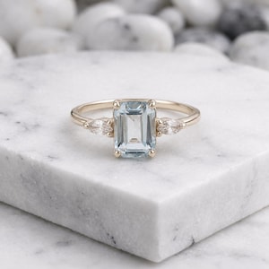 May include: A gold ring featuring a central emerald-cut light blue gemstone flanked by two smaller marquise-cut clear stones. The band is thin and polished, holding the three stones in a prong setting, creating a balanced and symmetrical design.
