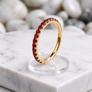 Könnte beinhalten: Ein dünner Goldring mit einer durchgehenden Reihe kleiner, runder, tiefroter Rubine, die entlang der Außenfläche gefasst sind. Der Ring steht aufrecht auf einem klaren, kreisförmigen Ständer und präsentiert die leuchtend roten Edelsteine.