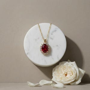 Könnte beinhalten: Eine Goldkette mit einem oval geschliffenen, tiefroten Edelsteinanhänger, der von einem Kranz aus kleinen, klaren Steinen umgeben ist. Der Anhänger hängt an einer zarten Goldkette und bildet ein klassisches Schmuckstück mit leuchtend roter Mitte.