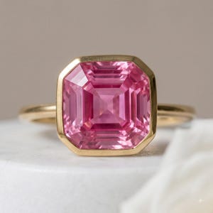 May include: A gold ring featuring a large, square-cut pink gemstone set in a smooth, polished gold bezel. The gemstone displays a faceted surface with deep pink hues and a symmetrical geometric pattern, mounted on a simple, slender gold band.