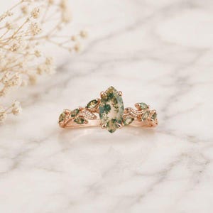 Könnte beinhalten: Ein Ring aus Roségold mit einem mittig platzierten, marquise-geschliffenen Moosachat. Die Ringschiene ist beidseitig mit blattförmigen Verzierungen gestaltet, die mit kleinen, klaren Steinen besetzt sind und sich entlang des Metalls erstrecken.